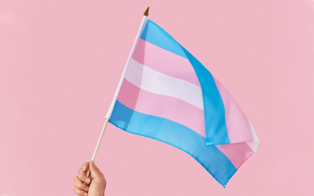 A person’s hand holds the transgender flag against a pink background.