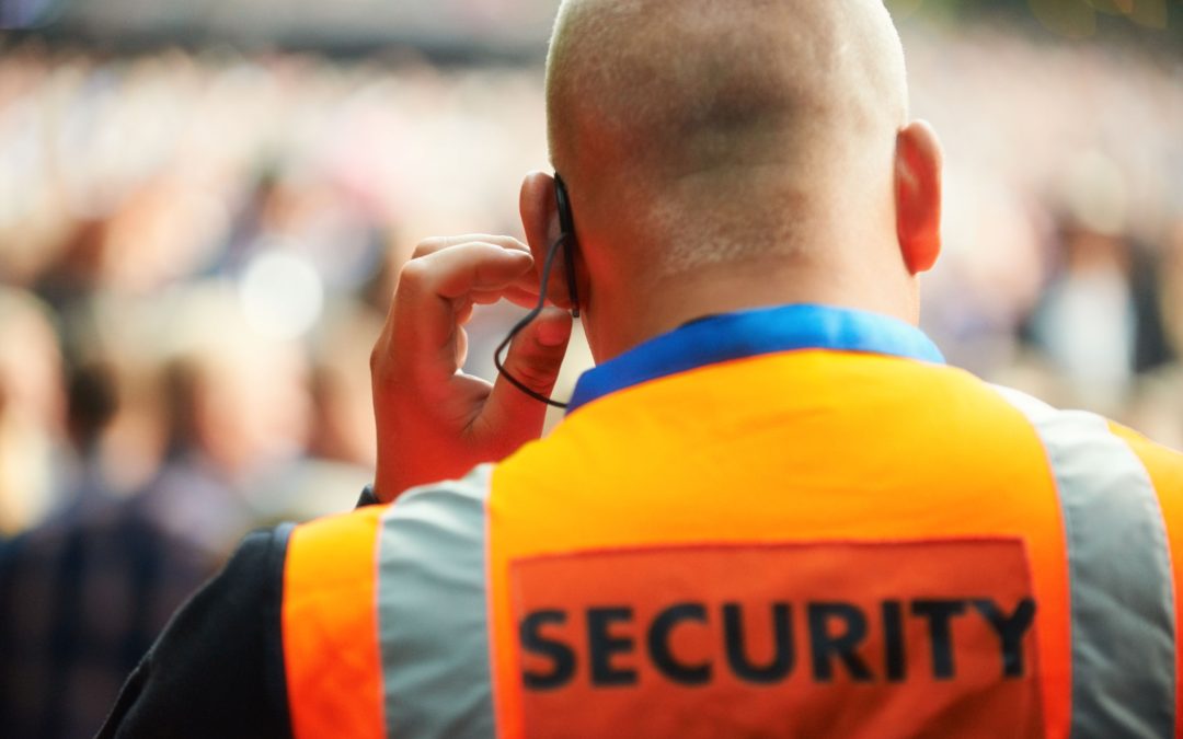 View of the back of a security guard's head in an orange vest, holding his hand to his ear piece as he controls a large crowd.