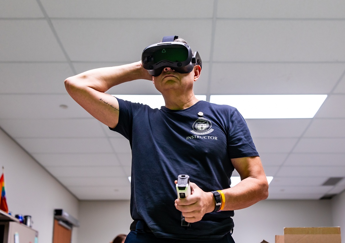 A male police instructor faces forward while wearing a virtual reality headset and holding a demo taser in his right hand. 